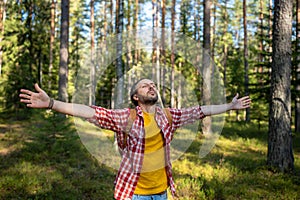 Pleased man stands in forest with outstretched arms, closing eyes in pleasure in harmony with nature