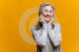 Pleased pretty woman holding hands near her face and smiling while posing against yellow studio wall. People and