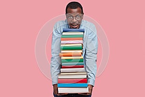 Pleased satisfied black male holds many books in hands, looks positively at camera, dressed in formal shirt, wears