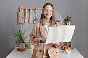 Pleased smiling woman freelancer with brown hair wearing beige jacket standing in kitchen interior working on laptop from home