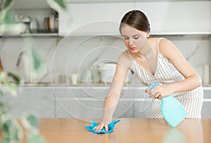 Positive young girl cleaning the surface of kitchen table