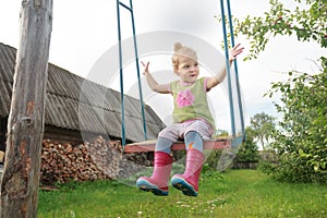 Pleased toddler girl wearing red kids gumboots riding on handmade rustic swing