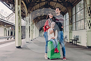 Pleased wife and husband cuddle with love, their daughter sits at suitcase, pose together on platform, look in ditance, wait for