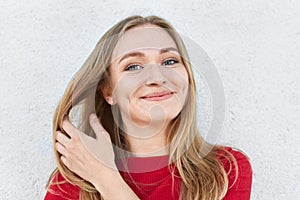 Pleased woman with blonde hair, luminous eyes and gentle smile having dimples on cheeks touching her beautiful hair with hand dres