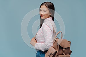 Pleased woman with dark hair, looks happily into distance, stands sideways with rucksack, going to travel, dressed in stylish