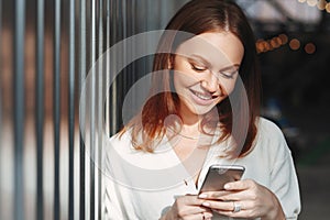 Pleased woman focused into screen of cell phone, checks email box, dressed in white clothes, sends feedback, connected to wireless