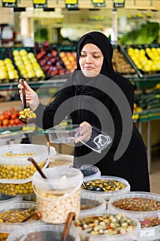 Pleased woman in sheila puts pickled olives into a container in the grocery section of supermarket