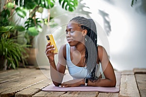 Pleased young african american woman lying on fitness mat with smartphone relaxing after training.