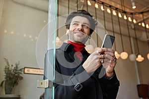 Pleased young handsome brown haired with earpieces keeping smartphone and looking cheerfully aside with wide smile, standing over