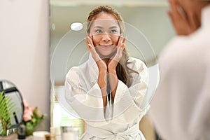 Pleased young woman wearing white bathrobe applying softening moisturizing face cream for skin face care in front of