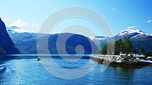 A pleasure boat is slowly leaving Valldal, Norway on the calm waters of a fjord on a sunny day with snow on the peaks.