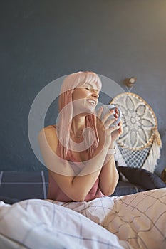 Pleasured lady with pink hair holds mug of delicious drink sitting on bed
