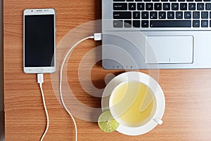 Plug in USB cord charger of the mobile phone with a laptop computer and freshly Lime juice in a white cup, on wooden floor