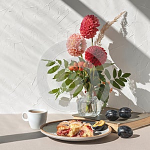 Plum pie piece on plate, cup of tea, vase with autumn flower bouquet on table, aesthetic sunlight shadows on white wall background