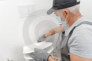 Plumber in medical mask installing toilet bowl in restroom, work in bathroom