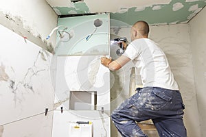 Plumber using electric screwdriver. Bathroom renovation concept. Marble ceramic tiles with spacers and grey cement walls in