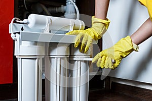 Plumber in yellow household gloves changes water filters. Repairman changing water filter cartridges in kitchen.