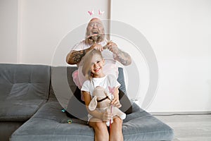 Plump daddy makes ponytail to daughter with fairy wings and toy dog in living room