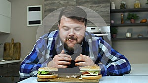 The plump guy enjoys his hamburger. The ketchup runs out of man`s fingers. The fat man with a beard bites a piece of