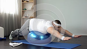 Plump male doing static exercise effort on fitness ball, desire to lose weight