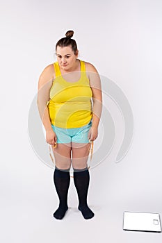 A plump woman in home clothes with a centimeter and floor scales, dissatisfied with her figure. Photo in full growth on