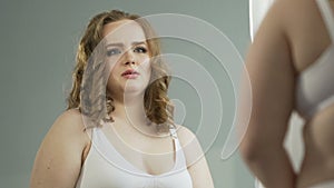 Plump woman in underwear studying carefully her reflection with sad look in eyes