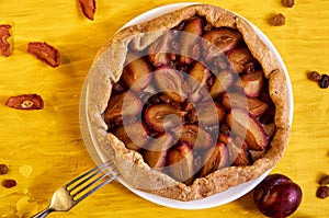 Plums pie with raisins on a white plate on yellow wooden background decorated with fresh plums, brown raisins