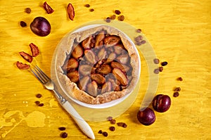 Plums pie with raisins on a white plate on yellow wooden background decorated with three fresh plums, brown raisins