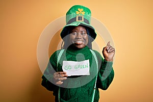 Plus size african american woman wearing green hat holding paper on saint patricks day surprised with an idea or question pointing