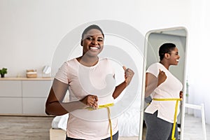 Plus size black woman measuring waist with tape in front of mirror, showing results of slimming diet, gesturing thumb up