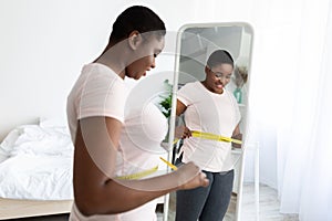 Plus size black woman measuring waist with tape, standing near mirror, satisfied with results of slimming diet at home