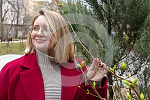 Plus size blonde in maroon coat enjoys communing with spring nature, first leaves and is happy for springtime. Close-up