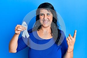 Plus size brunette woman holding blue ribbon smiling with an idea or question pointing finger with happy face, number one