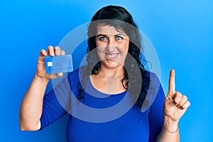 Plus size brunette woman holding credit card smiling with an idea or question pointing finger with happy face, number one