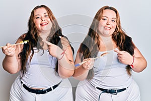 Plus size caucasian sisters woman eating eel sushi using chopsticks smiling happy pointing with hand and finger