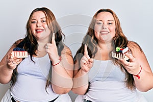 Plus size caucasian sisters woman holding slice of cake smiling with an idea or question pointing finger with happy face, number