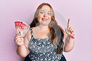 Plus size caucasian young woman holding 20 israel shekels banknotes smiling with an idea or question pointing finger with happy