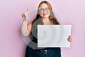 Plus size caucasian young woman holding blank empty banner smiling with an idea or question pointing finger with happy face,