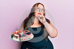 Plus size caucasian young woman holding cake slices serious face thinking about question with hand on chin, thoughtful about