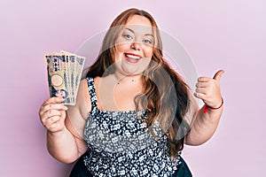 Plus size caucasian young woman holding japanese yen banknotes pointing thumb up to the side smiling happy with open mouth