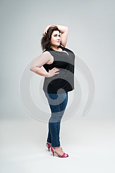 Plus size fashion model in black blouse and blue jeans, fat woman on gray background, body positive concept