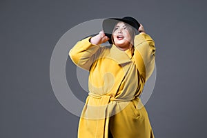 Plus size fashion model in yellow coat and black hat, fat woman on gray background