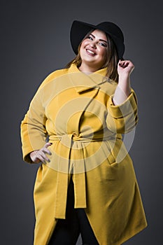 Plus size fashion model in yellow coat and black hat, fat woman on gray background, overweight female body