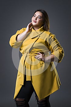 Plus size fashion model in yellow coat and black hat, fat woman on gray background, overweight female body