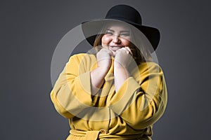 Plus size fashion model in yellow coat and black hat, fat woman on gray background, overweight female body