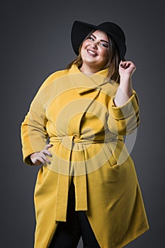 Plus size fashion model in yellow coat and black hat, fat woman on gray background, overweight female body photo