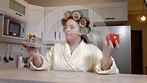 Plus size girl with curlers on her head dressed in a bathrobe chooses between an apple and a piece of cake