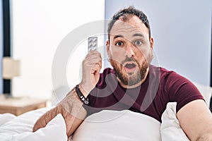 Plus size hispanic man with beard in the bed holding pills scared and amazed with open mouth for surprise, disbelief face