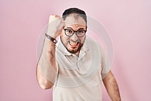 Plus size hispanic man with beard standing over pink background angry and mad raising fist frustrated and furious while shouting