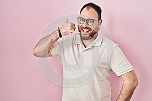 Plus size hispanic man with beard standing over pink background smiling doing phone gesture with hand and fingers like talking on